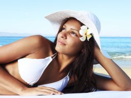 Woman in white bikini and sun hat relaxing by the ocean, symbolizing relaxation, wellness, and hormone balance in holistic RN-led health pro